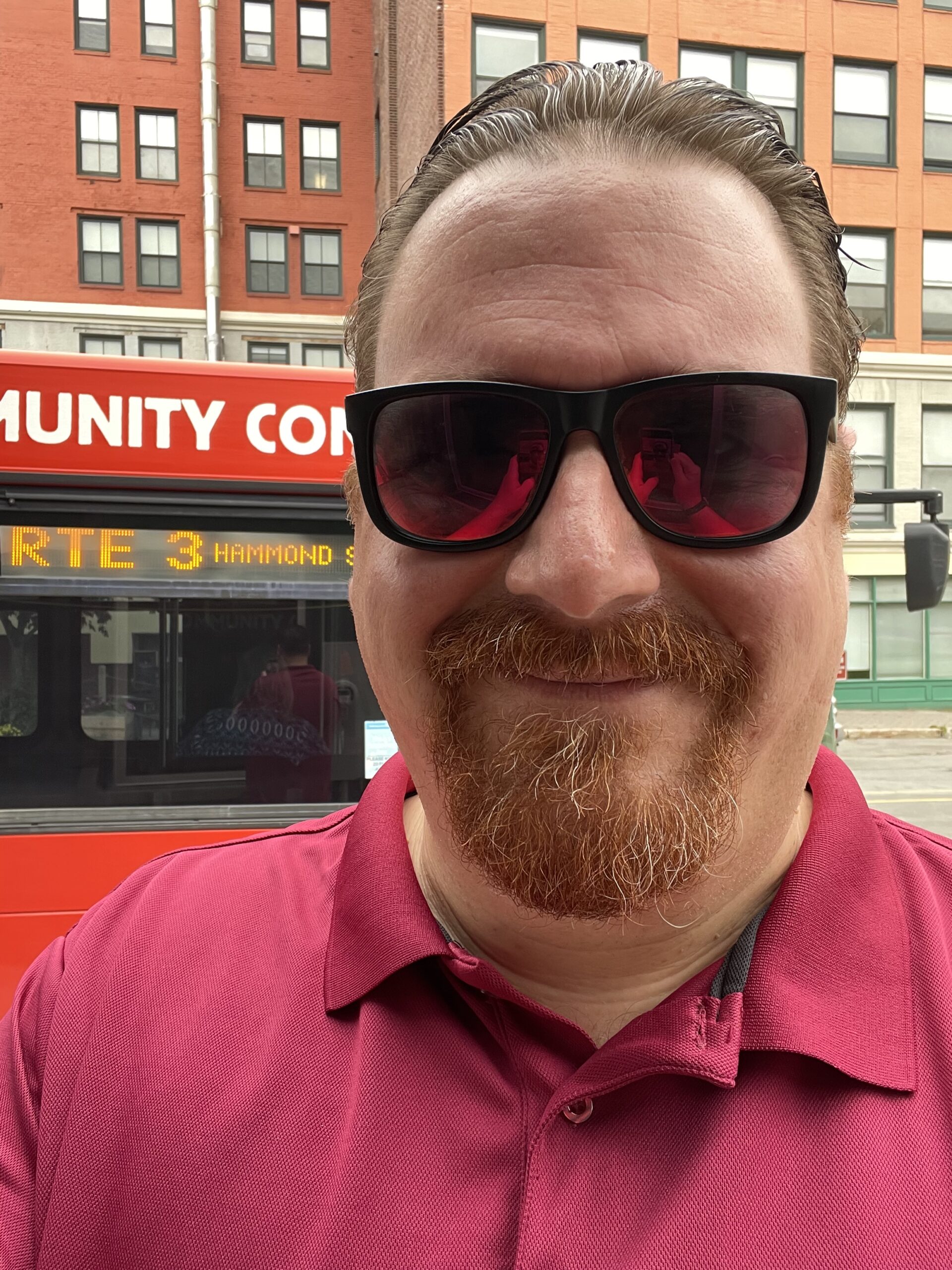 Michael Beck wearing sunglasses and standing in front of a Bangor city bus. It is a selfie. 